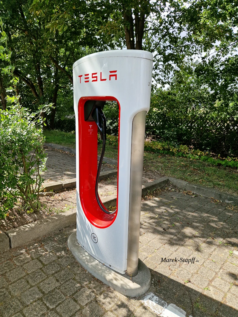 Tesla Supercharger V2 Ladesäule mit rotem Tesla-Logo auf weißem Mast vor blauem Himmel.